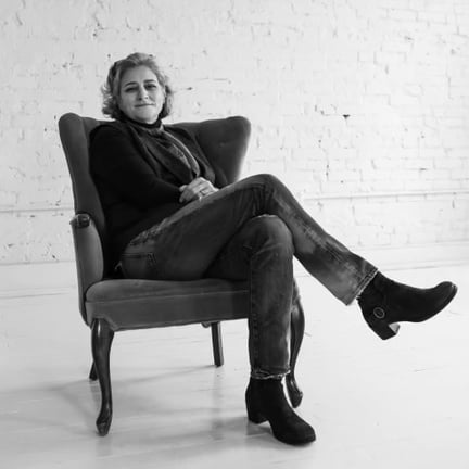 Black and white photo of Professor Dillard wearing a black swearer and jeans sitting back in a comfy armchair. Behind her is a white brick wall. 