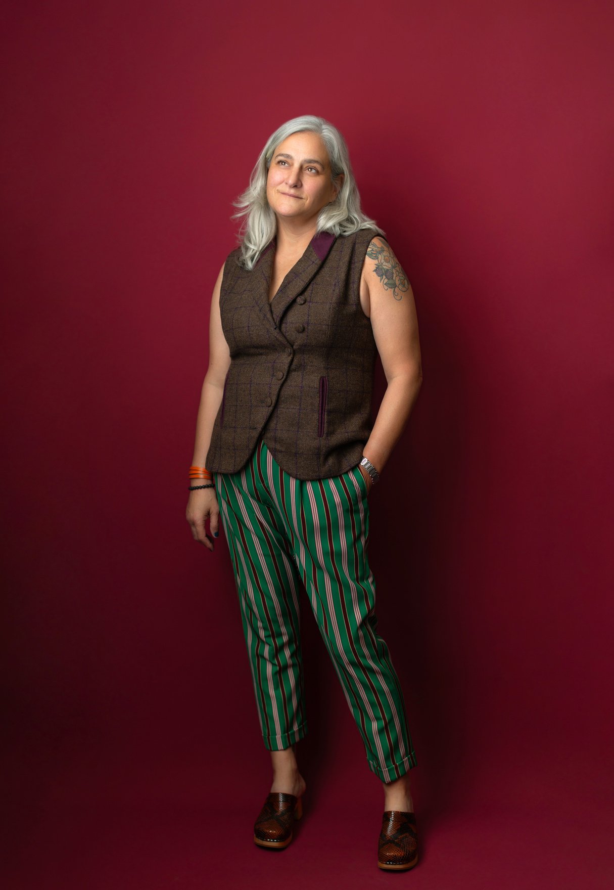 Professor Dillard with silver shoulder length hair standing in front of a burgundy red photo back drop. She has on striped pants and a vest. She is looking up towards the left corner of the photo. 